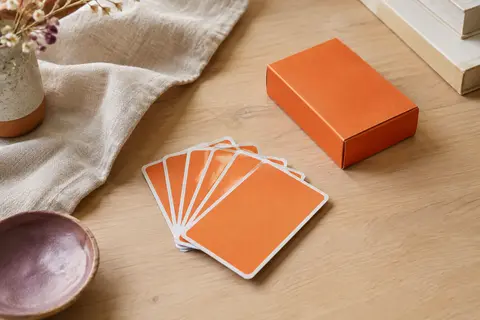 Jeu de cartes orange posé en éventail sur une table en bois clair avec du lin et de la céramique.