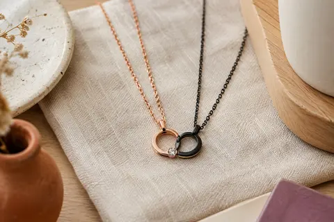 Deux colliers assortis aux pendentifs en anneau, l’un or rose avec zircon, l’autre noir, posés sur du lin.