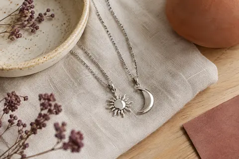 Deux colliers assortis avec pendentifs soleil et lune posés sur du lin clair près d’une coupelle en céramique.