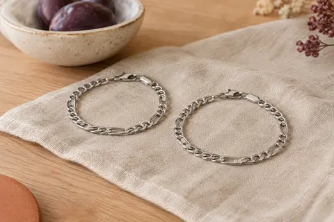 Deux bracelets assortis argentés à chaîne gourmette posés sur du lin dans un décor lumineux en bois clair.
