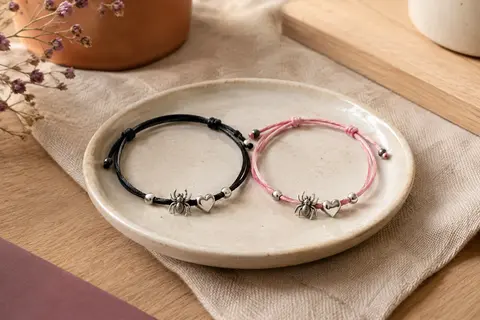Deux bracelets assortis en coton noir et rose, avec motif araignée et breloque cœur, posés sur de la céramique