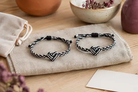 Deux bracelets assortis en nylon noir et blanc, avec nœud cœur, posés sur du lin près d’une pochette en velours.