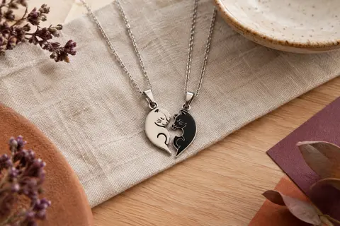 Deux colliers argentés et noirs, leurs pendentifs chats formant un cœur puzzle sur du lin.