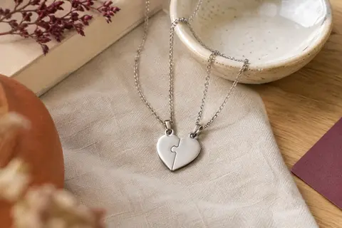 Deux colliers argentés avec pendentifs puzzle réunis en forme de cœur, posés sur du lin près d’une coupelle.