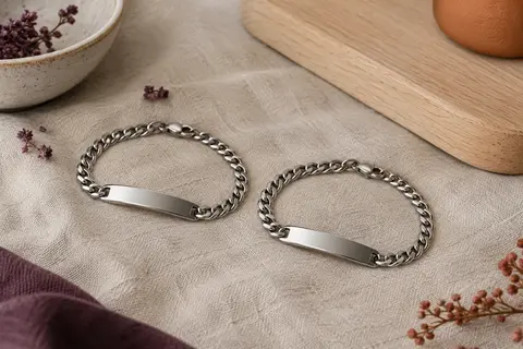 Deux bracelets gourmette argentés assortis, avec plaque lisse, posés sur du lin dans un décor clair.